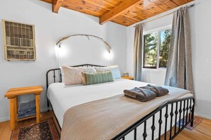 Un dormitorio con cama, mesita de noche, ventana con cortinas, unidad de aire acondicionado fijada a la pared y techo de madera.