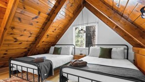 A loft bedroom with two beds, wooden beams, a window with blinds, and a hanging light fixture.