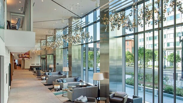 Eine moderne Hotelhalle mit einer langen Rezeption, mehreren Sitzbereichen mit Sofas und Sesseln, großen Fenstern mit Blick auf die Straße und dekorativen Hängeelementen.