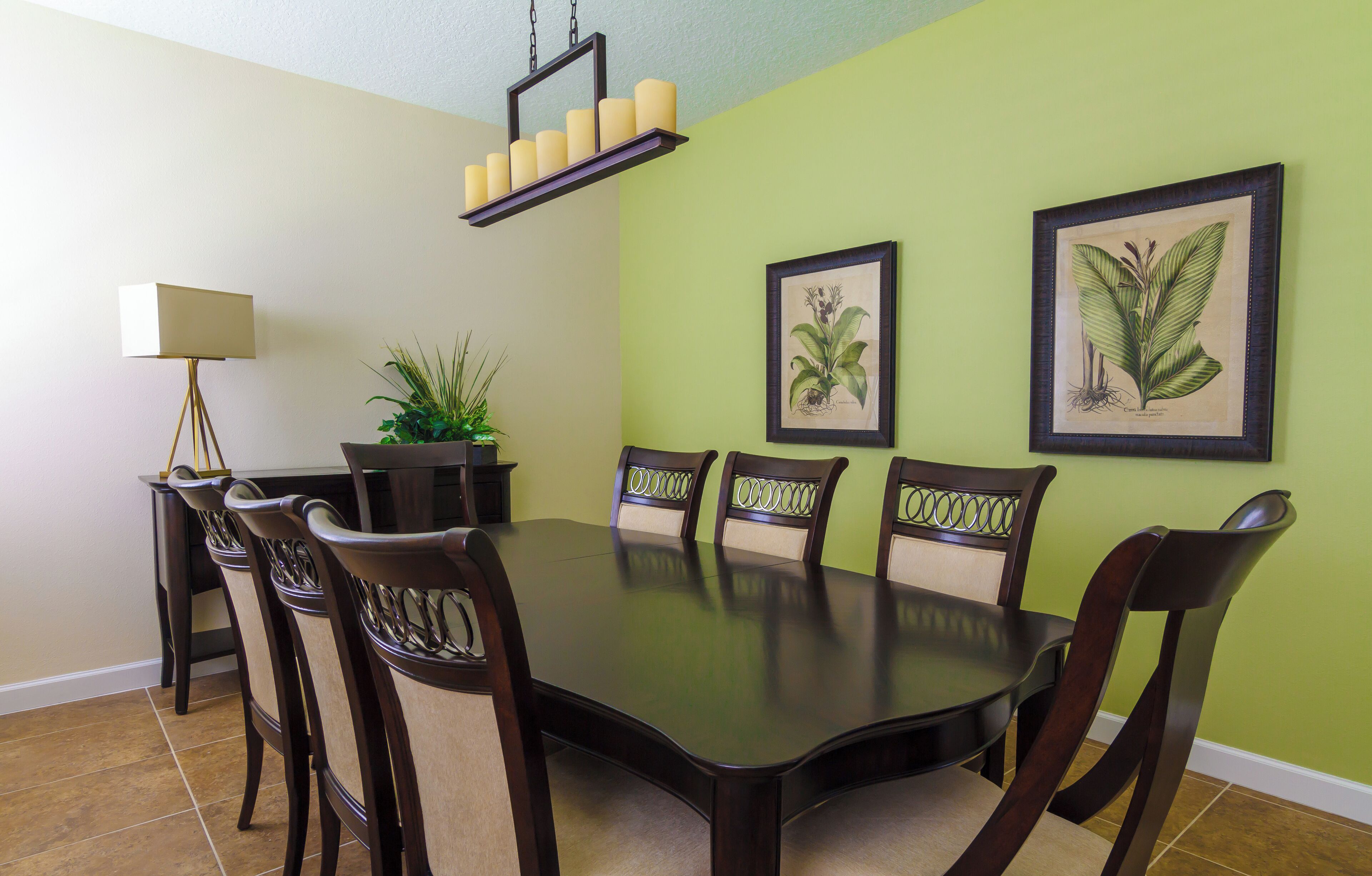 A dining area with a round table, six chairs, a floor lamp, and two framed botanical illustrations on the wall.