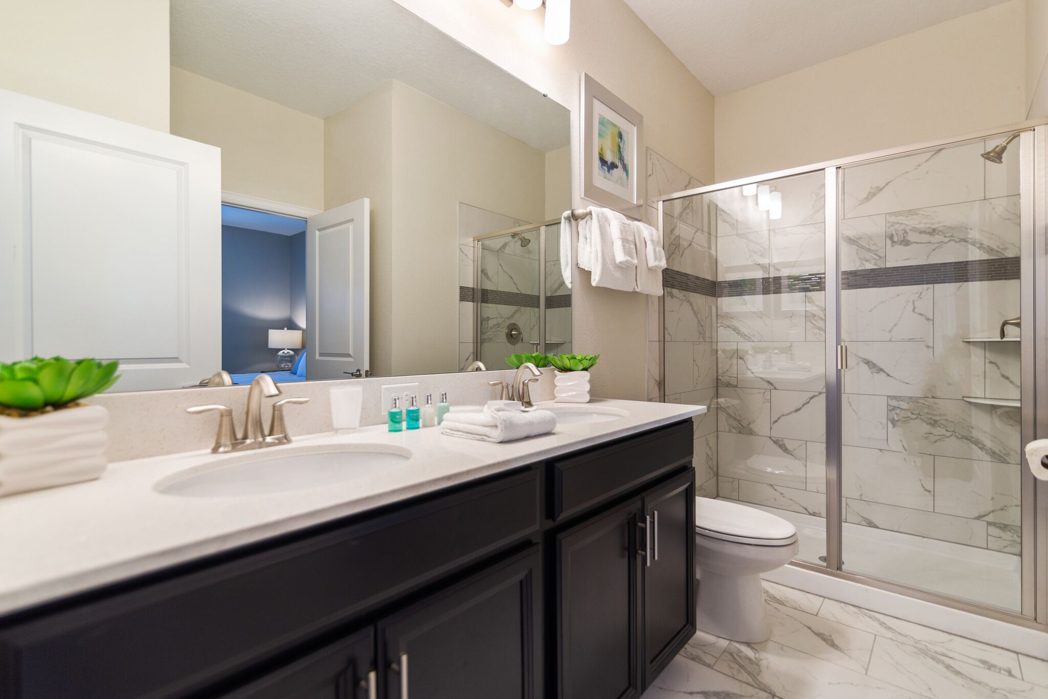 A modern bathroom with a large mirror, two sinks, a toilet, and a glass shower enclosure.