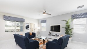 A modern living room with a glass coffee table, two blue sofas, a white entertainment unit, and a large TV. - 9038sid-the Retreat At Champions Gate 6 Bedroom Villa by RedAwning (Davenport)