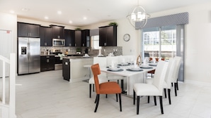 A modern kitchen with dark cabinets, stainless steel appliances, and a dining area with white chairs and a central island. - 9038sid-the Retreat At Champions Gate 6 Bedroom Villa by RedAwning (Davenport)