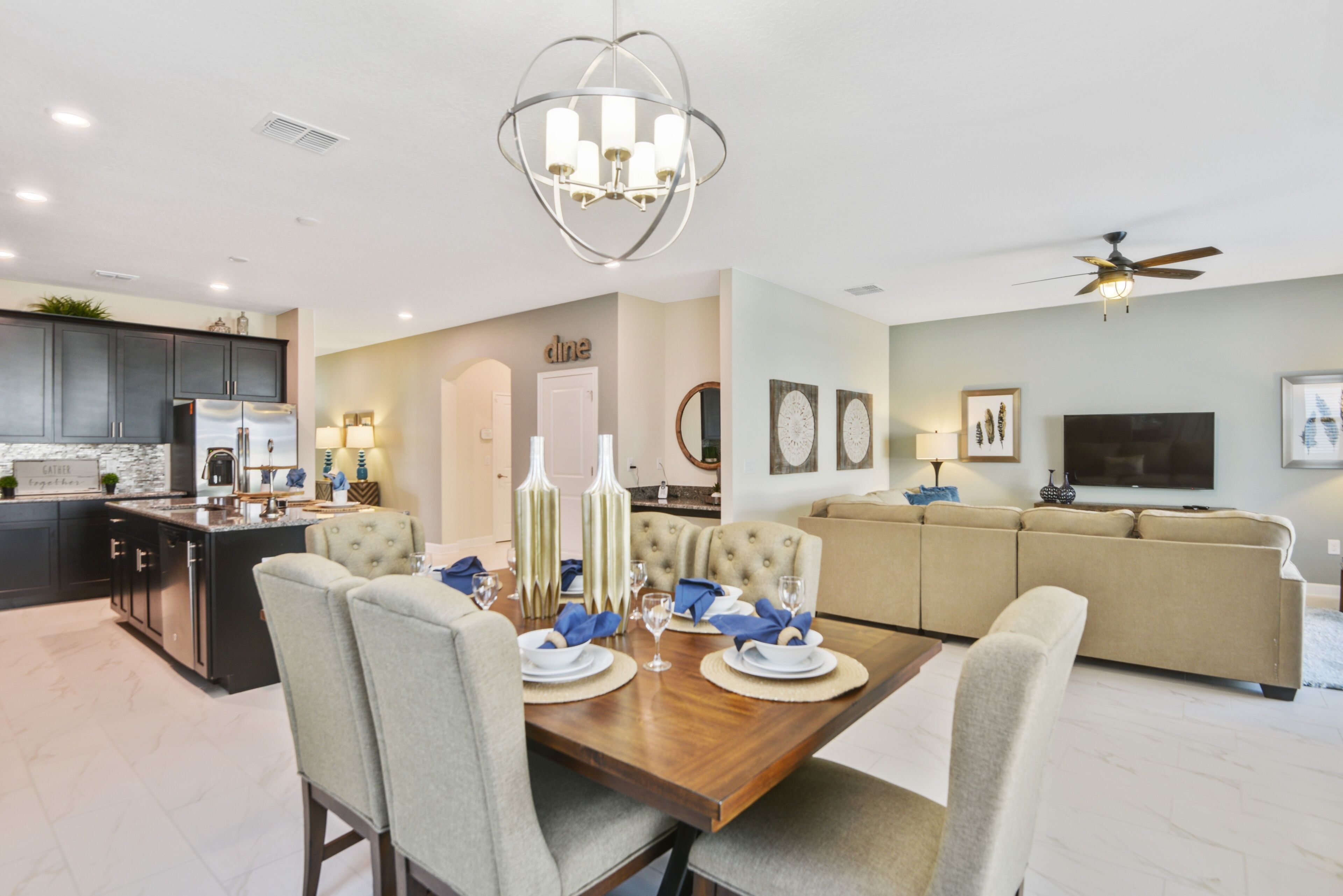 A modern kitchen with dark cabinets, a dining area with a wooden table set for four, and a living room with a beige sofa and a flat-screen TV.