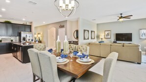 A modern kitchen with dark cabinets, a dining area with a wooden table set for four, and a living room with a beige sofa and a flat-screen TV.