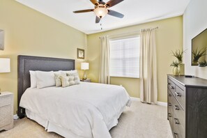 Une chambre à coucher avec un grand lit, un ventilateur de plafond, une table de chevet avec une lampe, une commode et une fenêtre avec des rideaux.