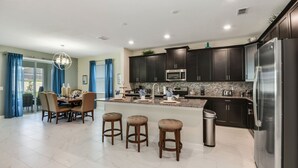 A modern kitchen with dark cabinets, a stainless steel refrigerator, a microwave, and a granite countertop. There are three bar stools and a dining table with chairs.