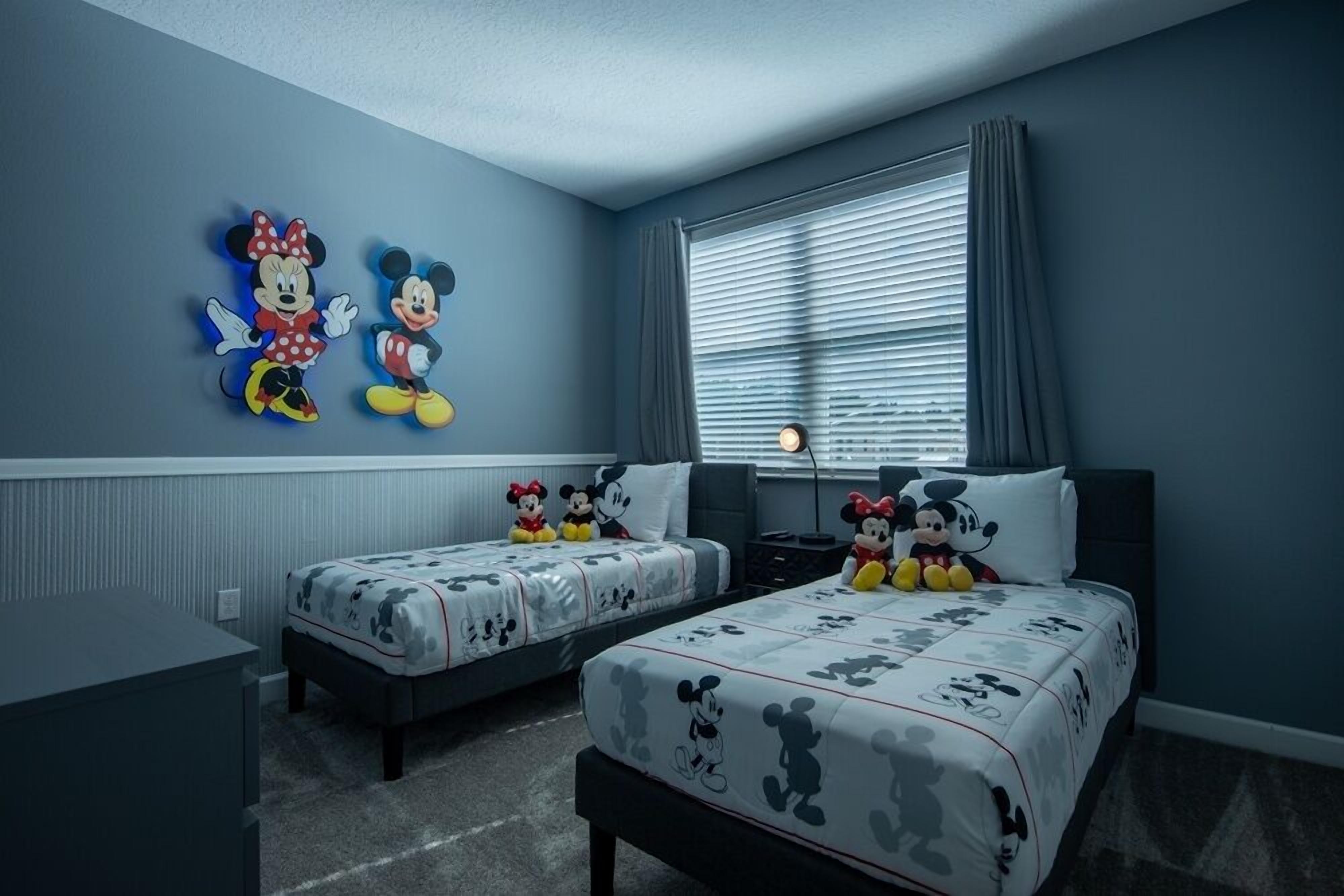 A bedroom with two beds, each with Mickey Mouse-themed bedding, a nightstand, a lamp, and a window with blinds.