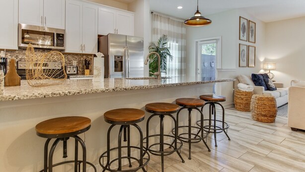 A modern kitchen with white cabinets, stainless steel appliances, and a granite countertop. There are four wooden stools at the bar area. - 1589mvd-the Retreat At Championsgate 4 Bedroom Townhouse by RedAwning (Davenport)