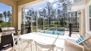 A covered outdoor area with a grill, a table, chairs, a pool, and a view of trees.