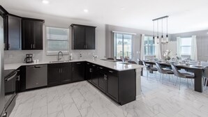 A modern kitchen with dark cabinetry, a large island, and a dining area with a table set for six. - 9028sh-the Retreat At Championsgate 6 Bedroom Home by RedAwning (Davenport)