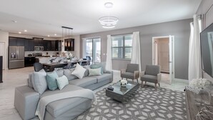 A modern living room with a grey sofa, armchairs, and a coffee table. There is a kitchen in the background with dark cabinets and stainless steel appliances. - 9028sh-the Retreat At Championsgate 6 Bedroom Home by RedAwning (Davenport)