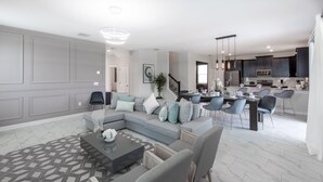 A modern living room with a sectional sofa, a dining area with chairs, and a kitchen with dark cabinets and stainless steel appliances. - 9028sh-the Retreat At Championsgate 6 Bedroom Home by RedAwning (Davenport)