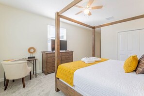 A four-poster bed with a white and yellow bedspread, a wooden desk with a computer, a chair, and a wardrobe.