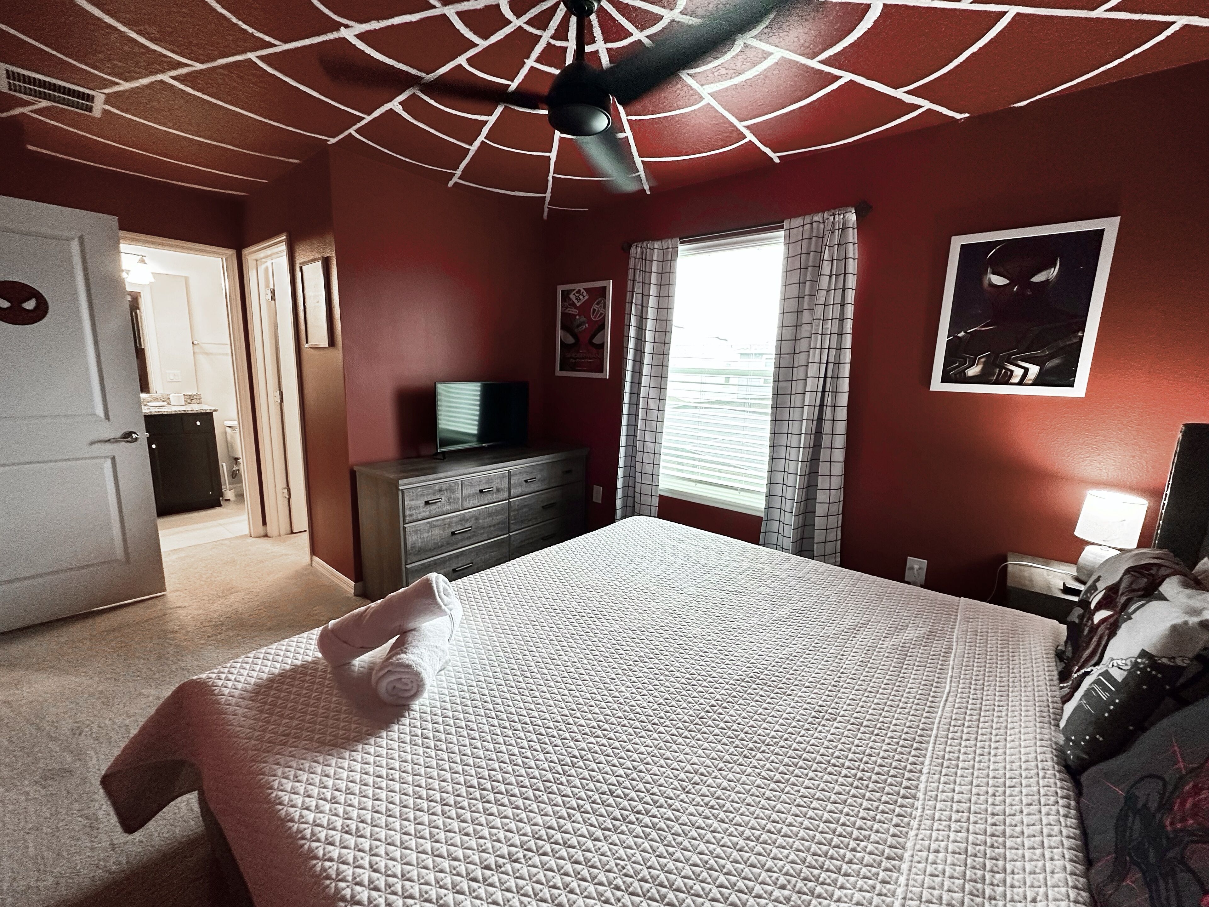 A bedroom with a bed, a dresser, a television on top, a window with curtains, and a ceiling fan.