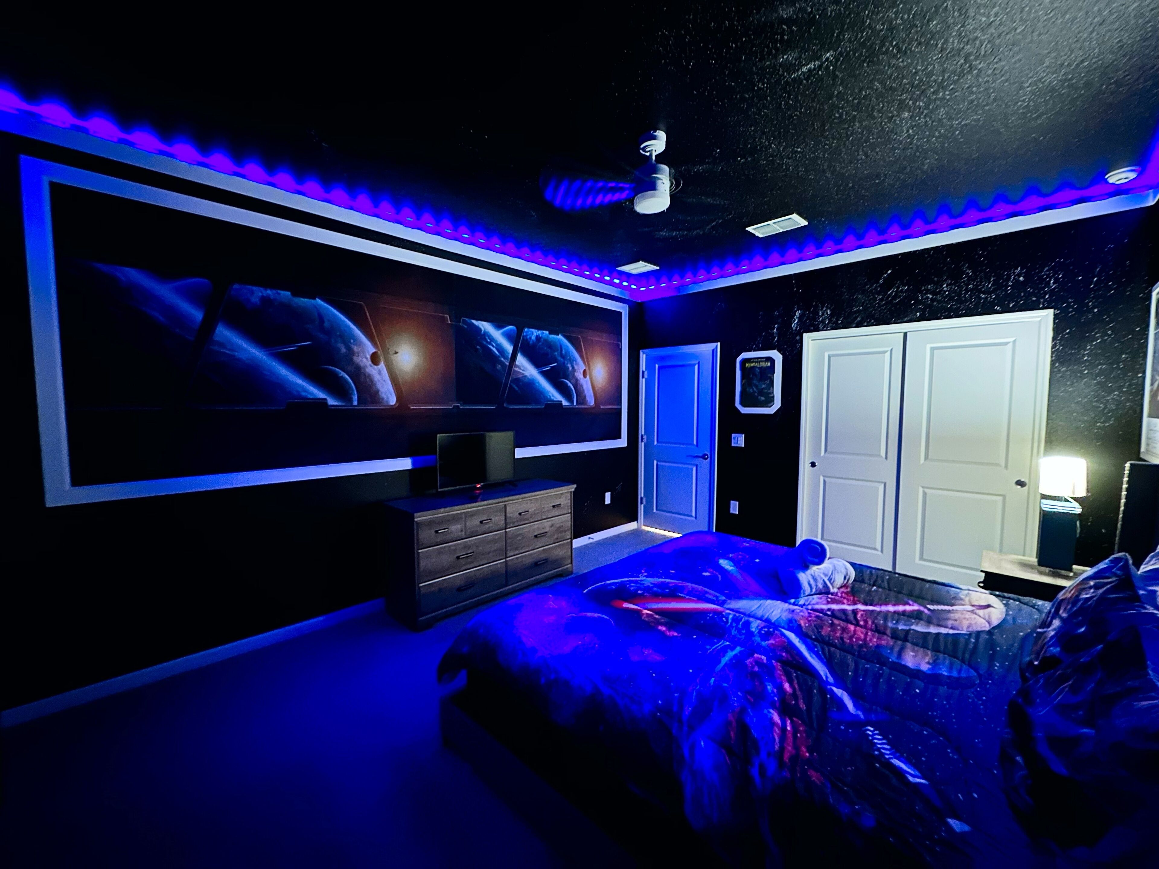A bedroom with a large bed, a dresser, a ceiling fan, and a wall-mounted TV displaying space-themed images.