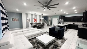A modern living room with a sectional sofa, a dark armchair, a coffee table, and a dining area with a table and chairs. - 2127lc- Windsor Island 10 Bedroom Home by RedAwning (Davenport)