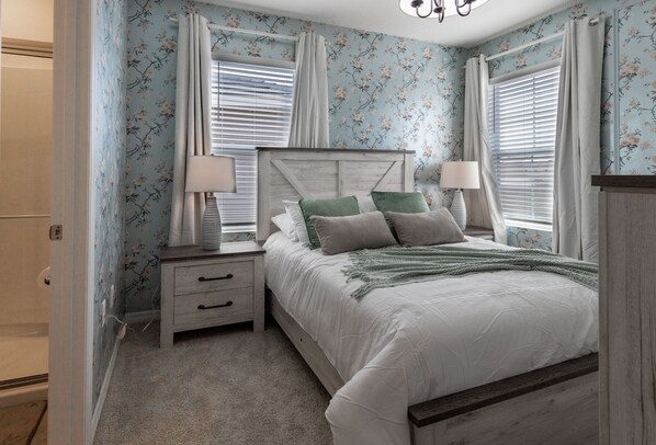 Un dormitorio con papel tapiz floral, cabecera de madera, cama con ropa blanca, dos mesitas de noche con lámparas y una ventana con persianas.