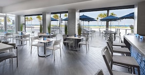 Ein modernes Restaurant mit Außenbereich, das auf den Strand blickt, mit weißen Tischen und Stühlen sowie blauen Sonnenschirmen.
