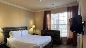 Una habitación de hotel con cama, tocador, televisor y ventana con cortinas.