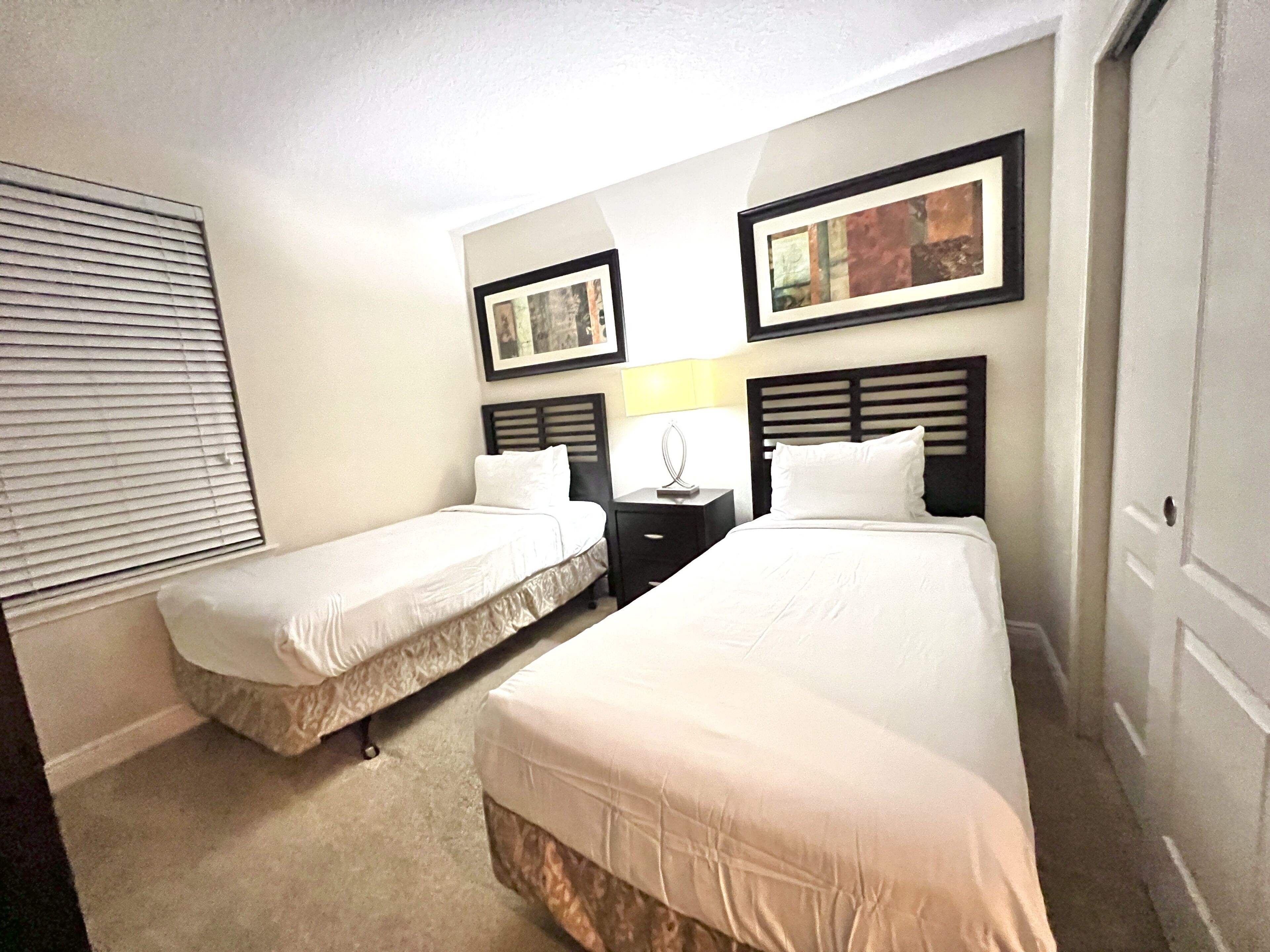 A hotel room with two single beds, a nightstand with a lamp, and two framed pictures on the wall.