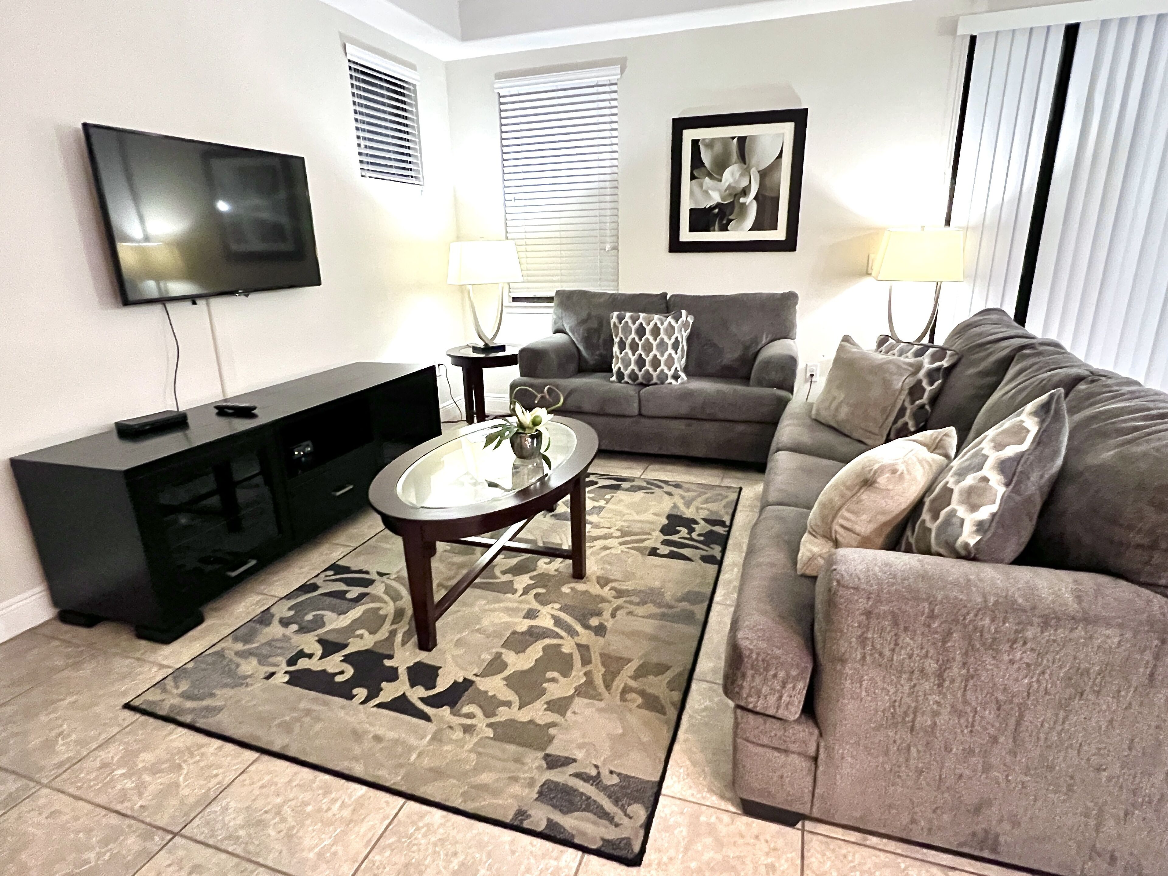 A living room with a flat-screen TV, a black entertainment unit, a grey sofa with patterned cushions, a coffee table, and a patterned rug.