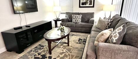 A living room with a flat-screen TV, a black entertainment unit, a grey sofa with patterned cushions, a coffee table, and a patterned rug.
