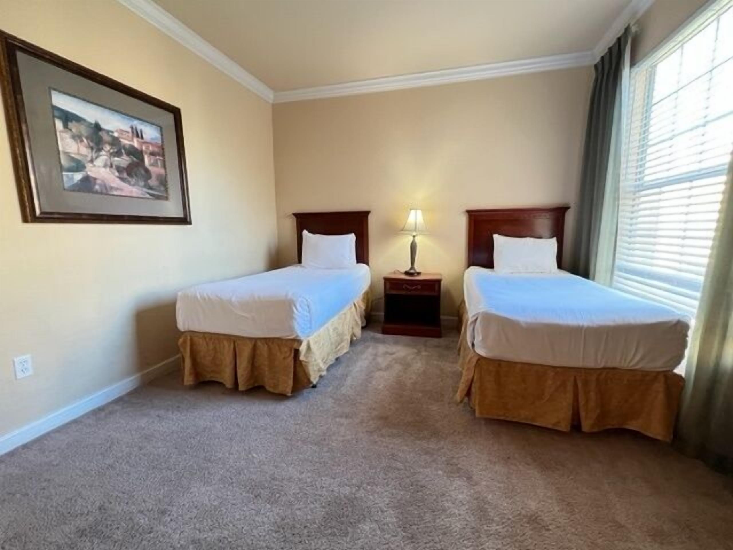 A hotel room with two single beds, a nightstand with a lamp, a framed picture on the wall, and a window with curtains.