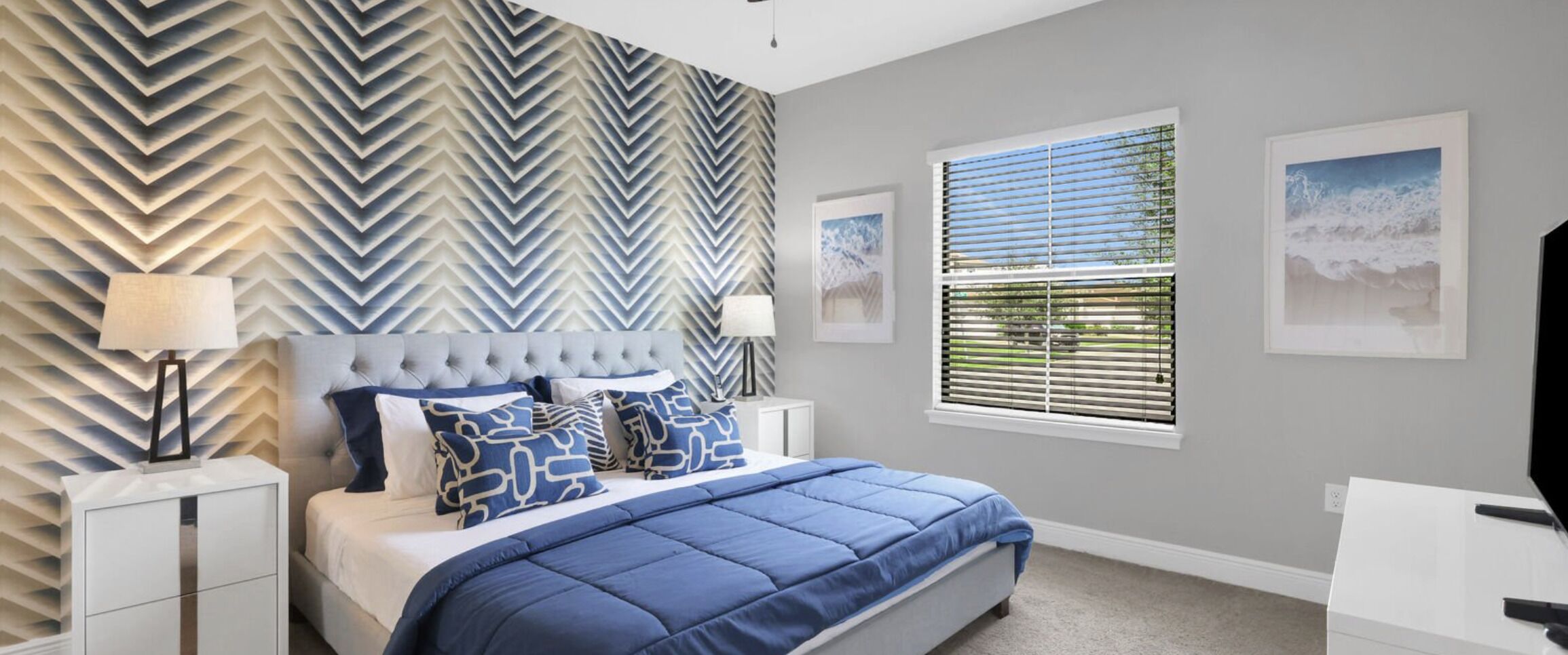 A bedroom with a chevron-patterned wall, a tufted headboard, a large bed with blue bedding, two bedside lamps, and a window with blinds.