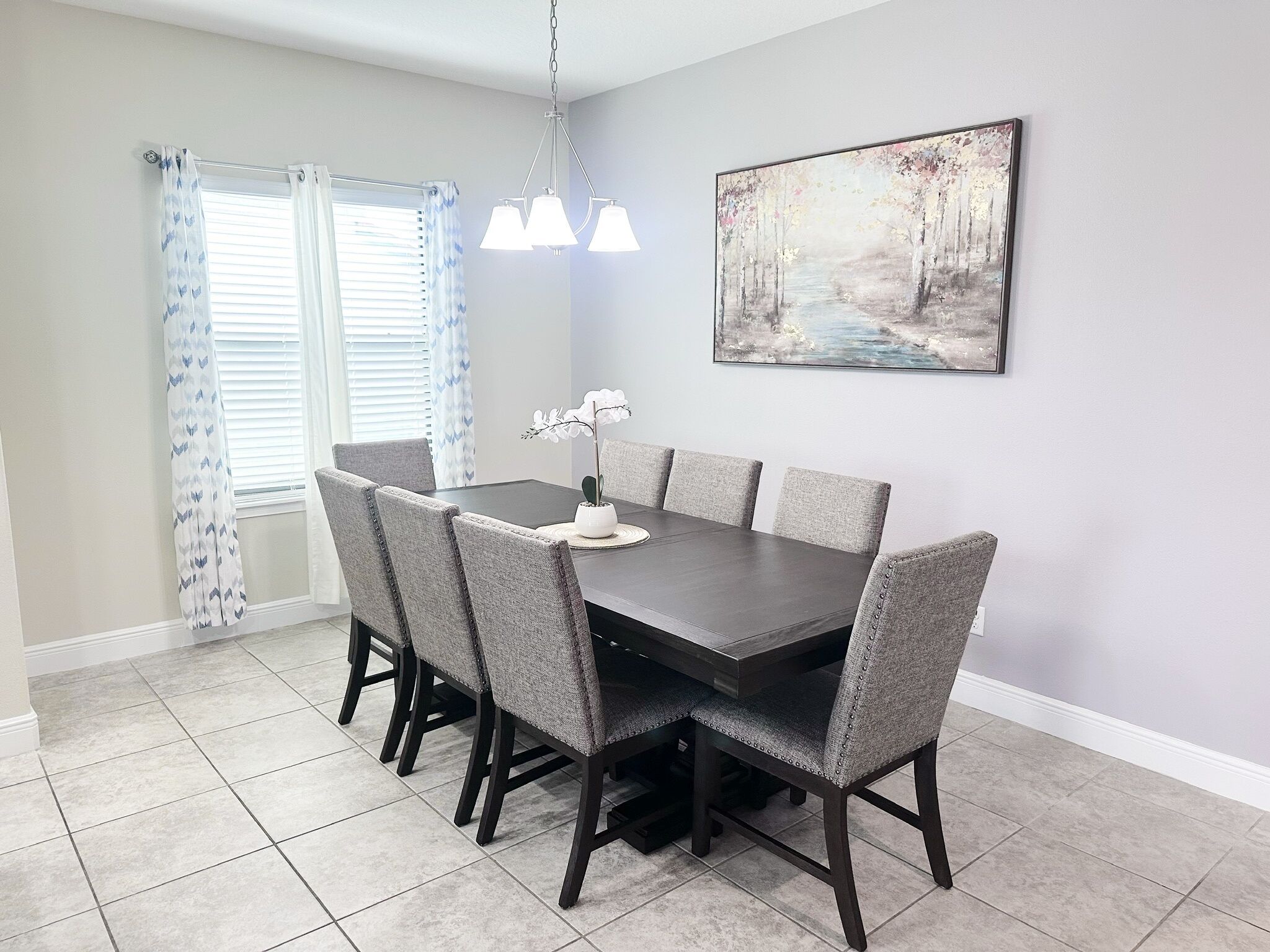 A dining room with a dark wooden table, grey upholstered chairs, a white vase with flowers, and a painting on the wall.