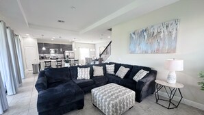 Ein modernes Wohnzimmer mit einem Ecksofa, einem Hocker und einem runden Beistelltisch mit Lampe. Eine Treppe führt auf eine obere Ebene, und im Hintergrund ist eine Küche zu sehen.