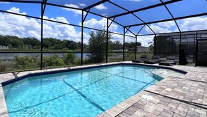 A swimming pool with a clear blue water, surrounded by a covered patio area with lounge chairs and a view of a lake and trees. - Balmoral Resort-140aa 5 Bedroom Home by RedAwning (Haines City)