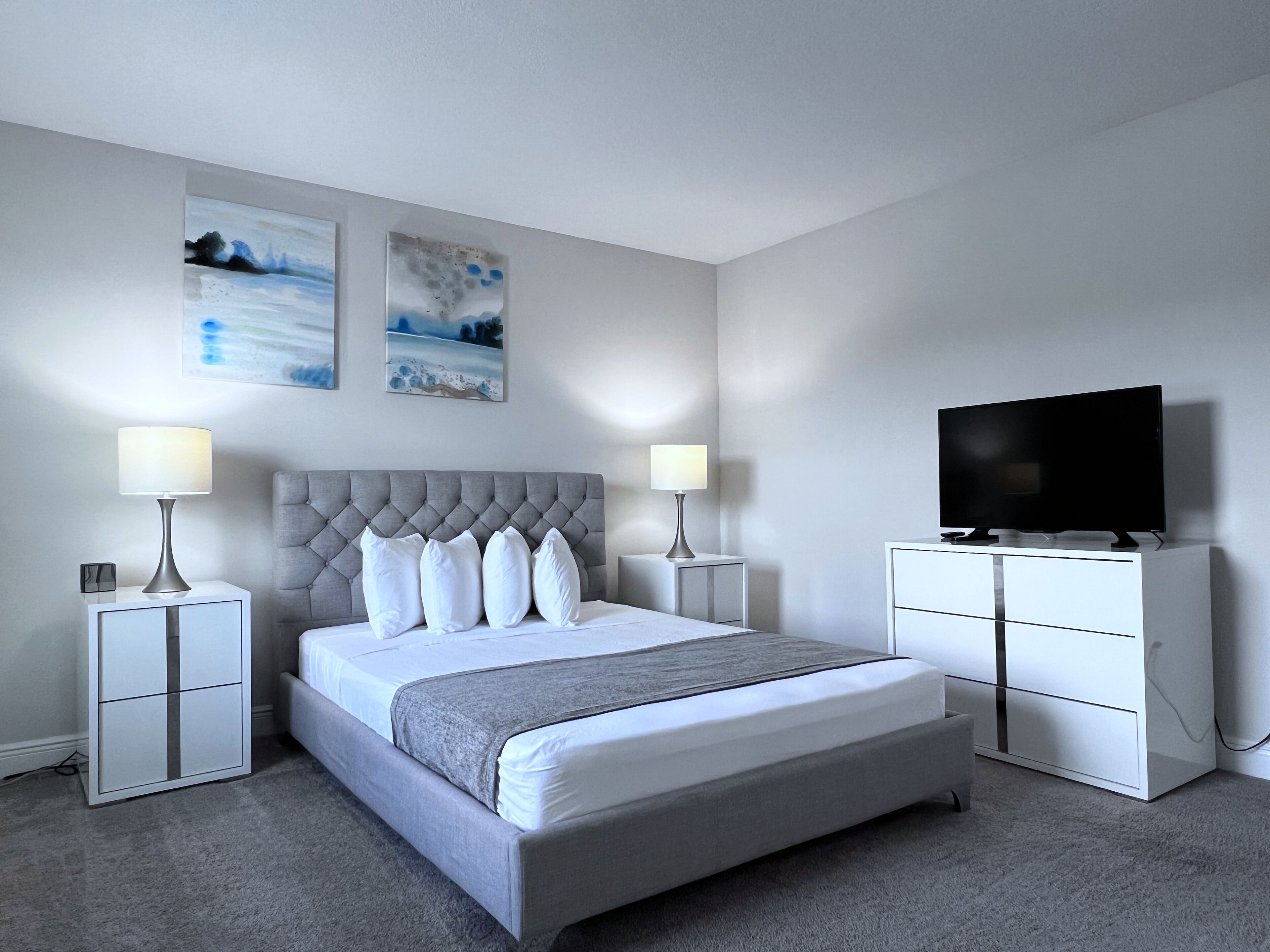 A modern bedroom with a tufted headboard, a large bed with white linens, two bedside tables with lamps, a flat-screen TV on a cabinet, and two abstract wall art pieces.