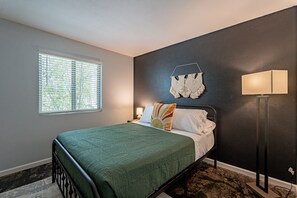 A bedroom with a bed, a nightstand, a lamp, a window with blinds, and a wall-mounted decoration.