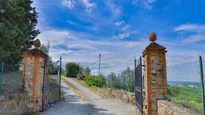A gated entrance with stone pillars and metal gates, leading to a pathway surrounded by greenery and a clear sky. - Villa in Certaldo ID 4125 (Certaldo)