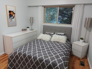 Quarto com cama, cómoda, mesa de cabeceira, candeeiro e janela com cortinas.
