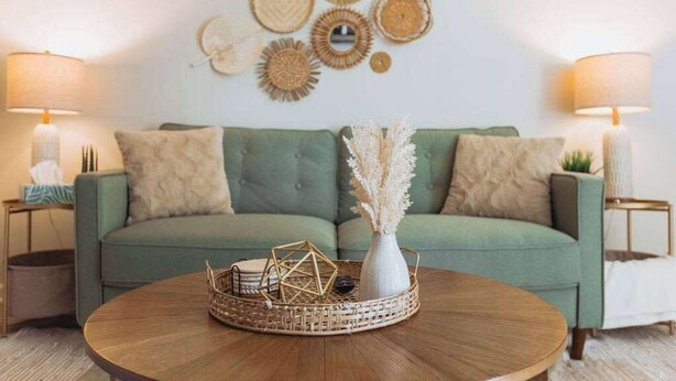 Un salon avec un canapé vert, deux tables d’appoint avec des lampes, une table basse avec un plateau en osier et des éléments de décoration murale.