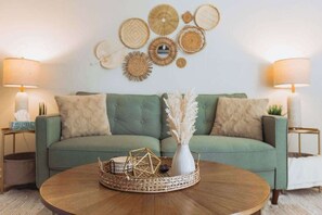 Sala de estar com um sofá verde, duas mesas laterais com luminárias, uma mesa de centro com bandeja trançada e detalhes decorativos nas paredes.