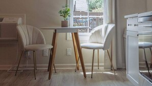 Une pièce comprenant une table à manger, deux chaises, une plante en pot, une fenêtre avec des rideaux et un coin cuisine.