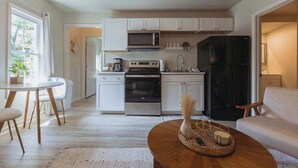 Une cuisine moderne avec des meubles blancs, un réfrigérateur noir, un four à micro-ondes et une cuisinière. On y trouve un coin repas avec une table ronde et des chaises, ainsi qu’un coin salon avec un canapé et une table basse.