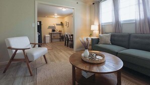 Una sala de estar con un sofá verde, un sillón blanco y una mesa de centro de madera con objetos decorativos. En el fondo, se observa un comedor con una mesa y sillas.