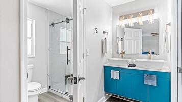 Une salle de bain moderne avec une douche à l’italienne en verre, des toilettes blanches, un meuble-lavabo bleu avec deux vasques et un grand miroir avec un éclairage situé au-dessus.