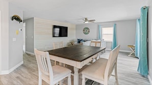 A dining area with a wooden table and white chairs, a ceiling fan, and a wall-mounted TV. - Golf Cart, 2 bed 2 Bath, Heated Pool (Indian Rocks Beach)