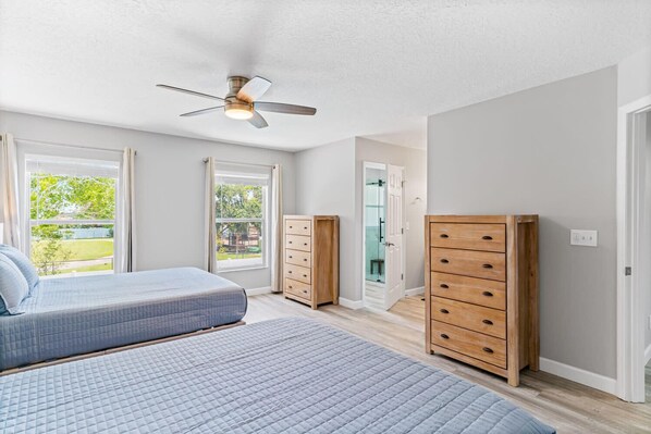 A bedroom with two beds, wooden dressers, a ceiling fan, and large windows with a view of the outdoors.