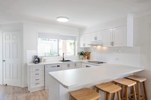 Une cuisine moderne avec des meubles blancs, un grand îlot central doté de tabourets, une fenêtre offrant une belle vue et un four encastré.