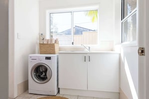 A laundry room with a washing machine, a sink, and a window with a view of a house and a fence.