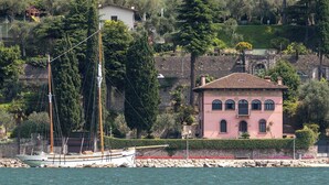 Un paysage côtier avec une villa rose, un voilier amarré près du rivage et une végétation luxuriante en arrière-plan.