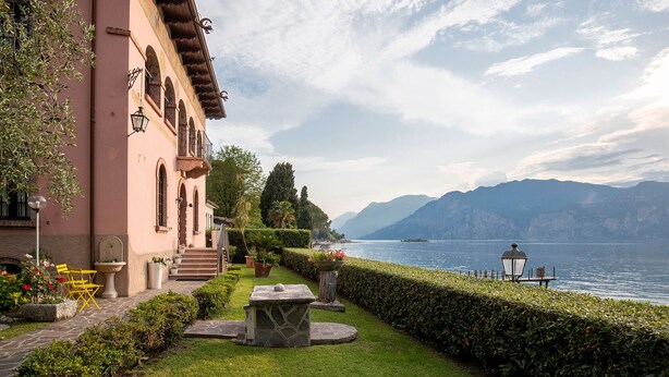Un bâtiment doté d’un balcon donnant sur un lac, entouré d’un jardin bien entretenu et offrant une vue dégagée sur les montagnes au loin.