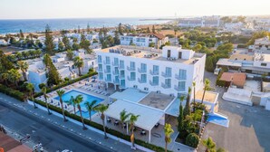 An aerial view of a modern hotel with a rooftop pool, surrounded by palm trees and a beach in the background.