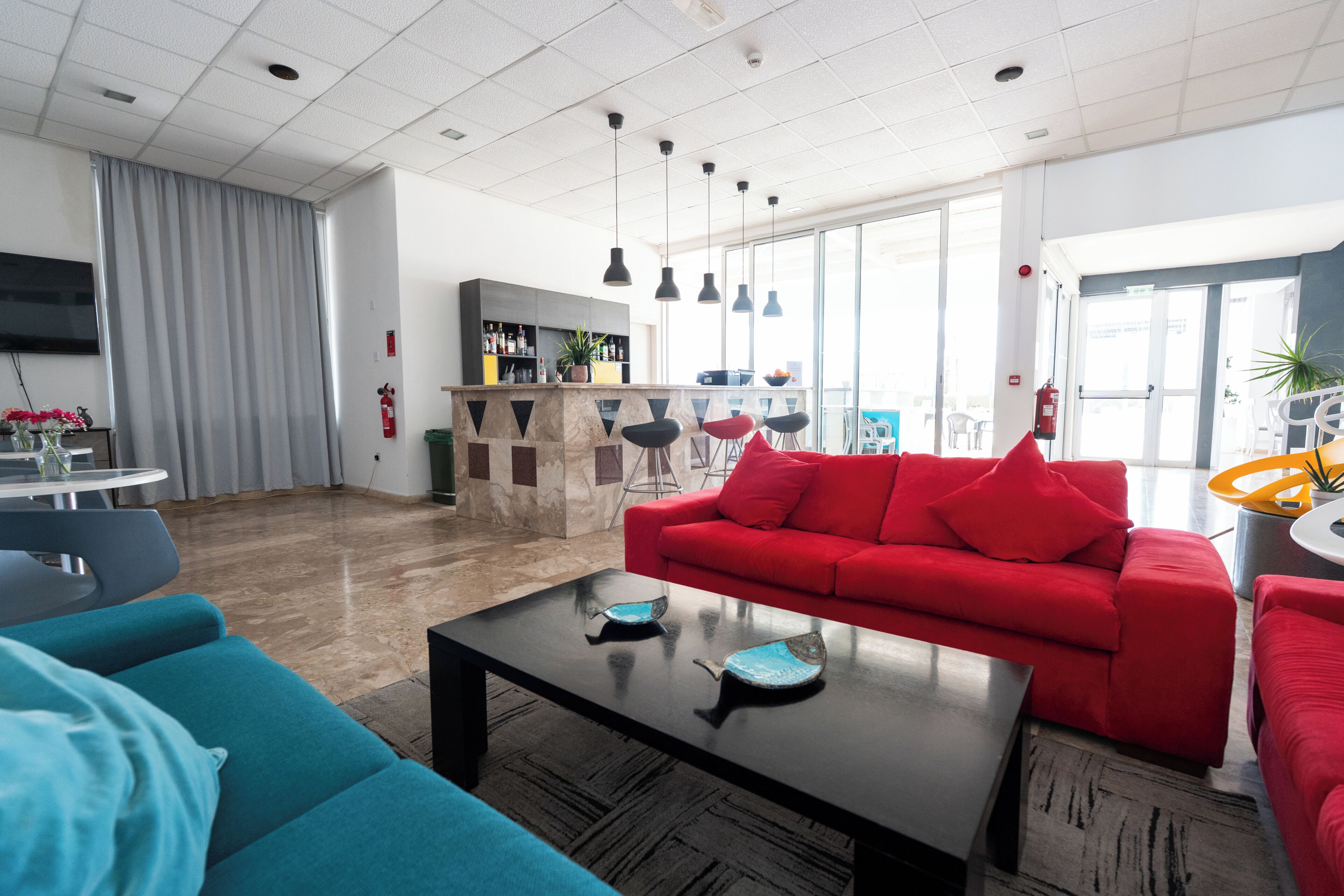 A modern hotel lobby with a red sofa, teal armchairs, a black coffee table, and a bar area with a large window view.